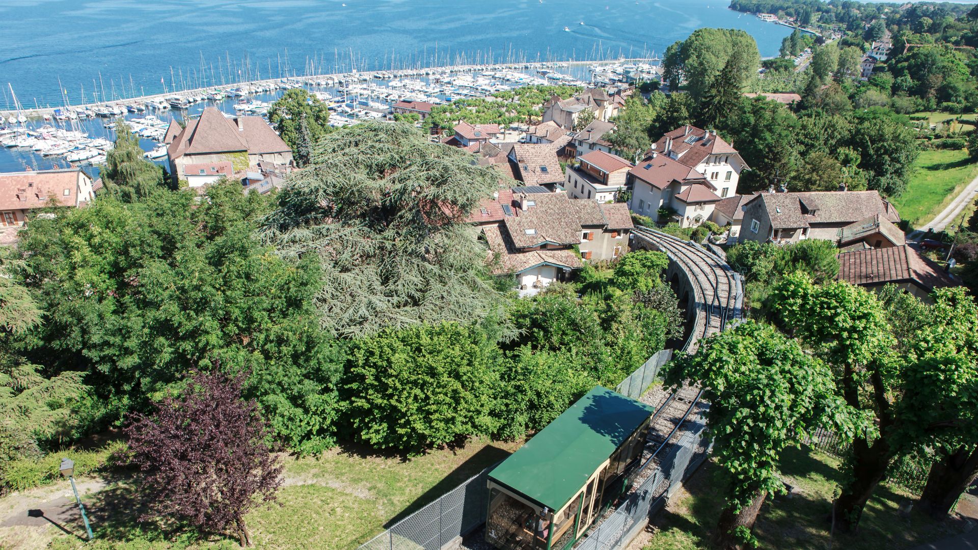 Vue aérienne sur un petit village avec des toits en tuiles et entouré de verdure, avec un funiculaire passant à travers et un port de plaisance en arrière-plan, situé à Thonon-les-Bains.