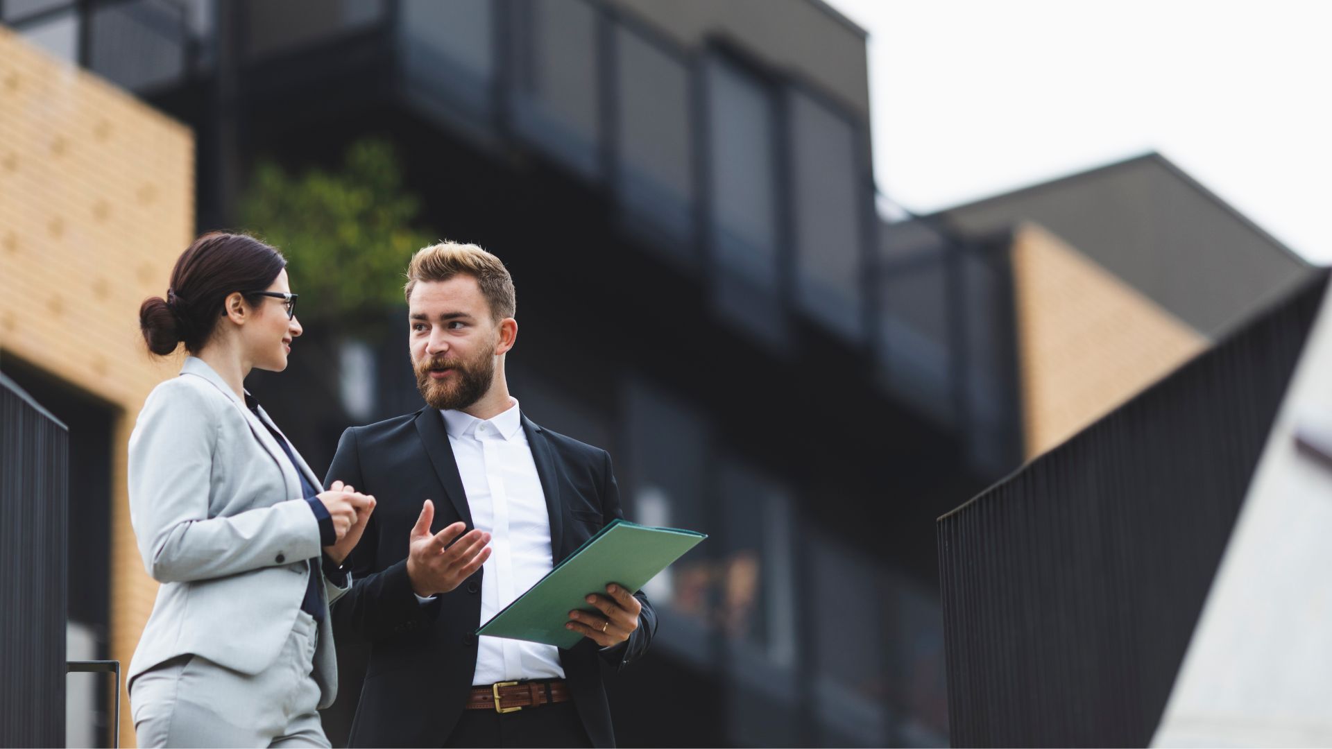 Un homme et une femme en tenue professionnelle discutant devant un bâtiment résidentiel moderne.